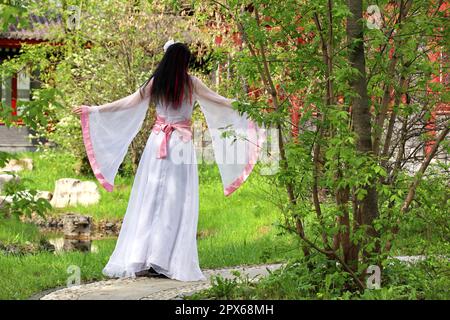 Girl in a traditional asian dress standing in garden on pagoda background, rear view. Cherry blossom season, asian beauty and culture Stock Photo