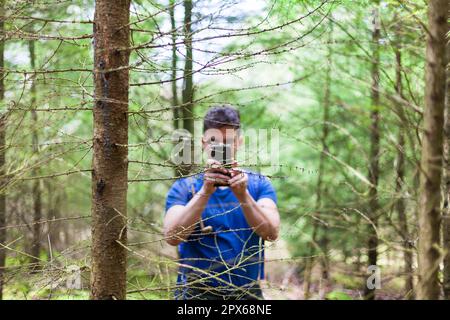 A young man is standing in the forest, holding a camera and taking a photo of himself. He is surrounded by lush trees and greenery Stock Photo