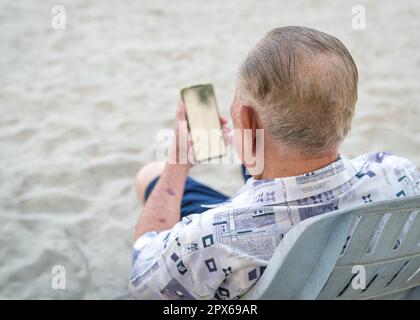 Back view of senior man using his smartphone to read. Digital lifestyle for the elderly. Stock Photo