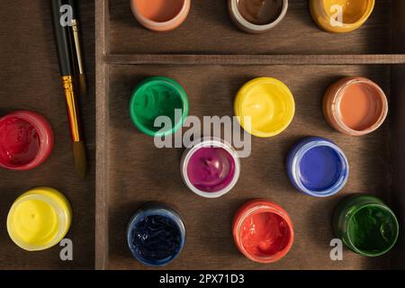 Gouache paints and paintbrushes on wooden easel, flat lay. Thirteen open bottles of artistic gouache paint and two synthetic paintbrushes in easel box Stock Photo