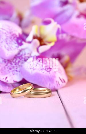 Golden wedding rings with purple orchids on pink wooden table. Vertical. Wedding ceremony, honeymoon, marriage proposal, Valentine's day in hot countr Stock Photo