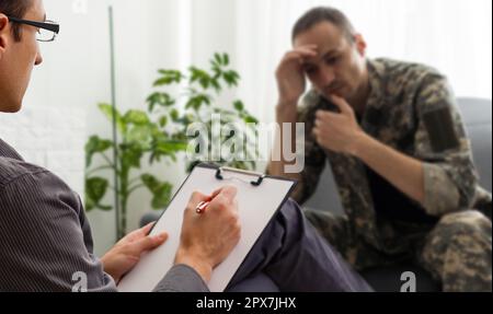 Psychologist making notes during therapy session with sad male soldier, PTSD Stock Photo