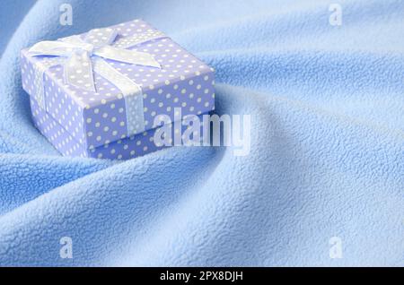 A small gift box in blue with a small bow lies on a blanket of soft and furry light blue fleece fabric with a lot of relief folds. Packing for a gift Stock Photo