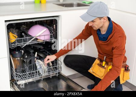 A man or a service worker in special clothes using a screwdriver attaches to the countertop or removes the dishwasher Stock Photo
