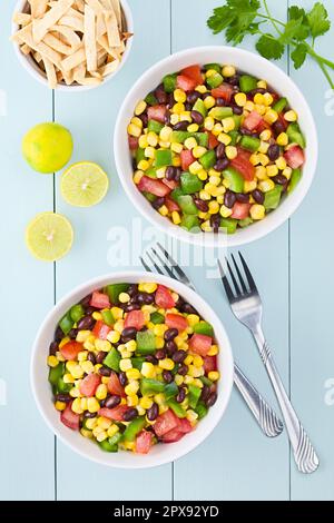 Mexican style colorful fresh vegetable salad made of beans, corn, tomato and bell pepper served in bowls, baked tortilla strips, limes and forks on th Stock Photo