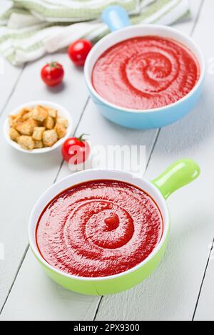 Fresh homemade tomato soup in colorful bowls, wholegrain croutons on the side, photographed on white wood (Selective Focus, Focus in the middle of the Stock Photo