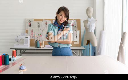 Young dressmaker in striped scarf holding scissor with a smile. Pin cushion, silk thread and ruler are on the workbench. Asian woman working in home b Stock Photo