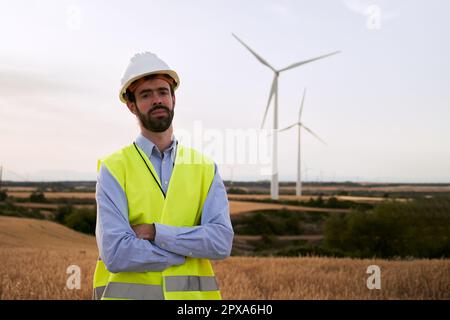 Technical engineer poses confidently with arms crossed looking at camera standing in a wind farm. Stock Photo