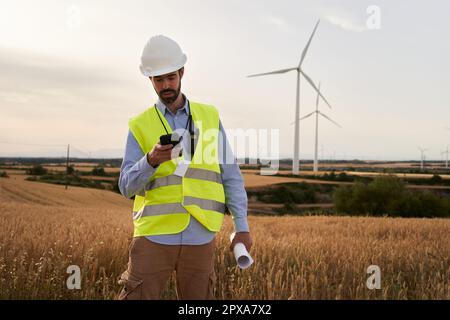 Boss engineer using mobile phone standing in wind farm. Worker looking at cell in renewable energy. Stock Photo