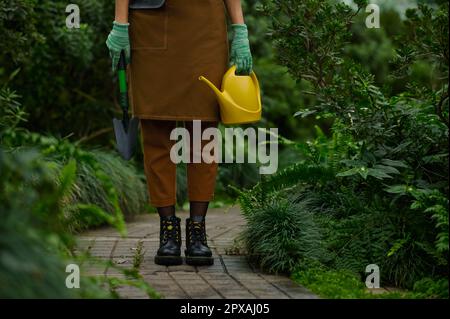 Cropped shot of female gardener dressed in apron holding garden tool in hands wearing rubber gloves view on leg in boots over greenhouse background Stock Photo
