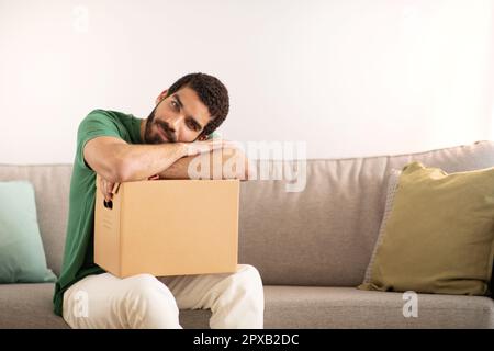 Tired serious pensive young middle eastern guy blogger resting on cardboard box, sits on sofa Stock Photo