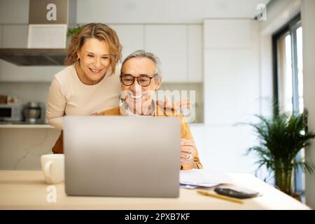 Happy senior spouses paying bills online from home, sitting in front of laptop with papers, kitchen interior, copy space Stock Photo