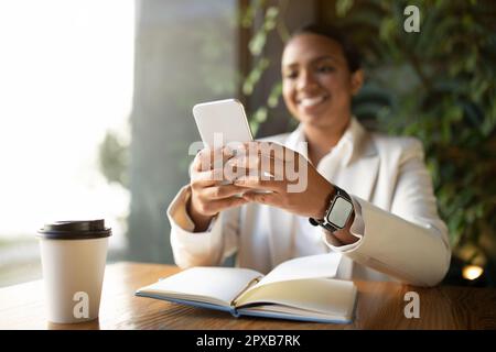 Positive young black lady in white suit at table chatting on phone, with cup of coffee and notebook in cafe Stock Photo