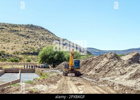 Boegoeberg Dam, South Africa - Feb 28 2023: A backhoe loader clearing sand that blocked the irrigation canal near Boegoeberg Dam. Workers are visible Stock Photo