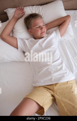 Calm young handsome teenager lies in bed in a white t-shirt in the bedroom at home and looks out the window. Rest, relaxation, good mood, lifestyle. M Stock Photo