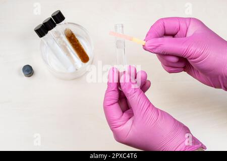 gloved hands holding test tube with liquid and indicator paper showing pink color, acid pH level over light desk Stock Photo