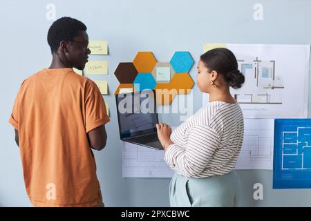 Couple of architects using laptop at their teamwork while examining blueprints on the wall in office Stock Photo