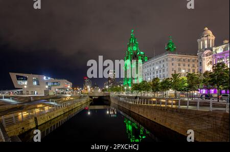 The Royal Liver Building lit up green at night alongside The Three Graces on Liverpool Waterfront. The ferry terminal and ship canal reflected. Stock Photo