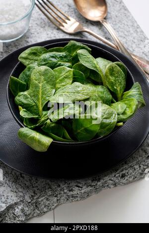 Fresh baby spinach in a black bowl and silver cutlery on a gray stone Stock Photo