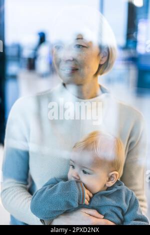 Thoughtful young mother looking trough window holding his infant baby boy child while waiting to board an airplane at airport terminal departure gates Stock Photo