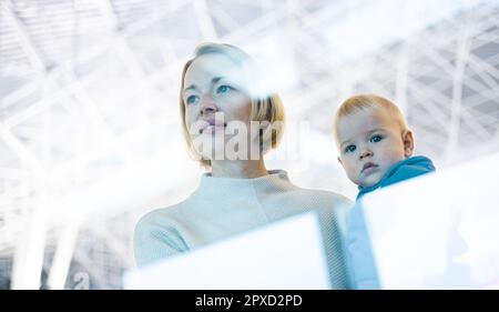 Thoughtful young mother looking trough window holding his infant baby boy child while waiting to board an airplane at airport terminal departure gates Stock Photo