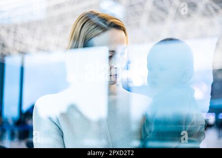 Thoughtful young mother looking trough window holding his infant baby boy child while waiting to board an airplane at airport terminal departure gates Stock Photo