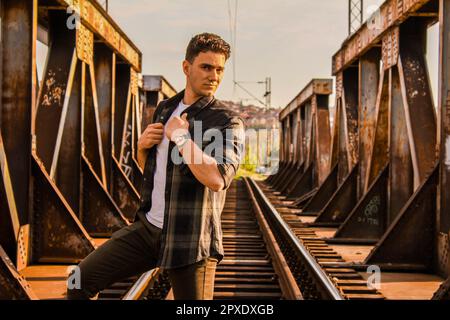 Caucasian teenager standing and watching the camera on train tracks on a rusty train bridge with a blurry city in the background Stock Photo