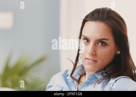 A close-up portrait of a young woman with brown hair and light-colored eyes, wearing a denim shirt and indoors. In the blurred background, a plant and Stock Photo