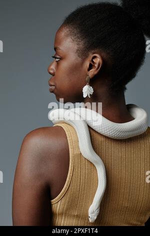 Rear view of young gorgeous African American woman in earrings posing with white rat snake enlacing her neck and creeping down back Stock Photo