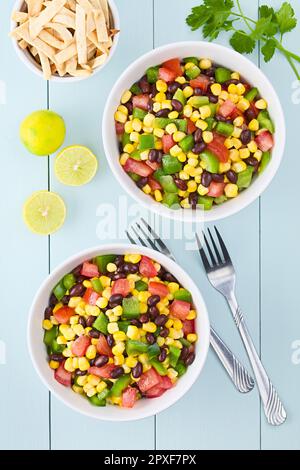 Mexican style colorful fresh vegetable salad made of beans, corn, tomato and bell pepper served in bowls, baked tortilla strips, limes and forks on th Stock Photo