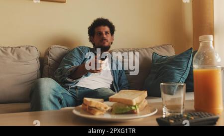 Guy dressed in denim shirt has lunch when he sits in the living room with remote control in his hands Stock Photo