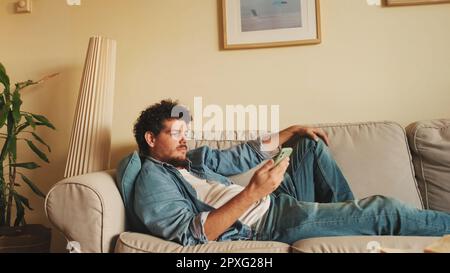 Happy guy holds smartphone in his hands, sends text message, communicates in social networks, surfs the web while sitting indoors Stock Photo