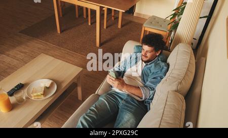 Happy guy holds smartphone in his hands, sends text message, communicates in social networks, surfs the web while sitting indoors Stock Photo