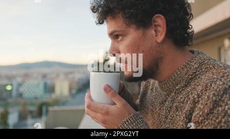 Peaceful guy looks at the city from the balcony and drinks coffee, enjoys the beautiful view Stock Photo