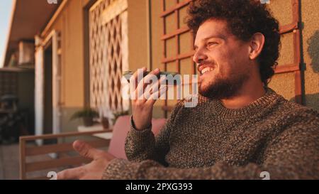 Happy guy sitting on the terrace and chatting on smartphone using speakerphone Stock Photo
