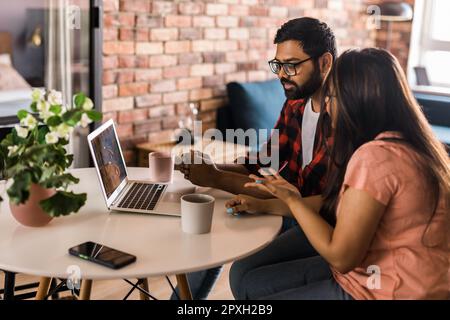Happy indian family couple cuddle at desk make video call to friends using laptop webcam. Loving young spouses look at computer screen waving hands in Stock Photo