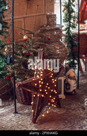 A Christmas tree with red small toys in a wicker pot, a snowman and star made of twigs and decorated with a garland, all standing on the paving stones Stock Photo