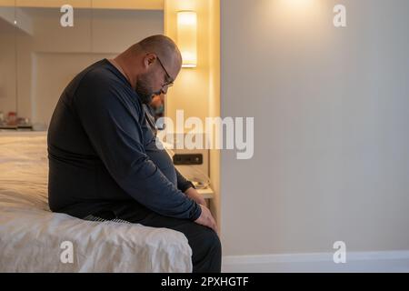 Overweight sad man resting alone in hotel having midlife crisis sitting on bed and boring. Stock Photo