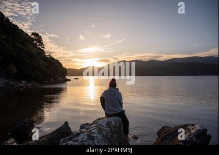 A woman sits in front of a sunset across a bay Stock Photo