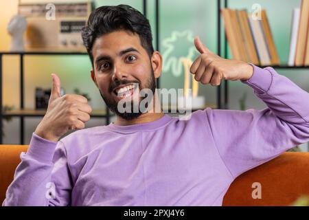 Like. Happy excited indian man looking approvingly at camera showing thumbs up, like sign positive something, good great news, positive feedback. Young hindu guy sitting on couch at home living room Stock Photo