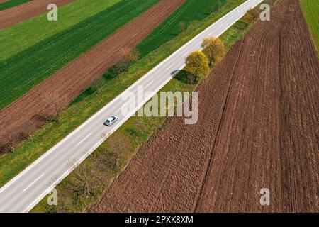 Aerial view of scenic countryside landscape in spring with country road and passenger car on it driving along on sunny spring day, drone pov. Transpor Stock Photo