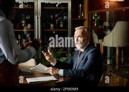 Mature businessman discussing menu together with waiter while sitting at table at lunch time in cafe Stock Photo
