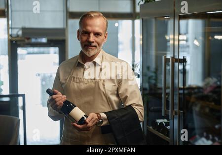 Portrait of mature waiter holding bottle of red wine and looking at camera while standing in restaurant Stock Photo