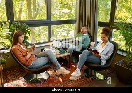 Young people business team meeting at eco friendly coworking space. Diverse millennial coworker sitting in circle, talking, holding documents and gadg Stock Photo