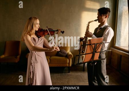 Young female violinist and male saxophonist training at home studio before music concert performance Stock Photo