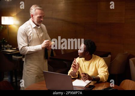 Mature waiter in uniform making notes while taking order from customer in restaurant Stock Photo