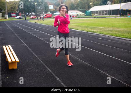 Young lady with a smartphone runs for fitness enjoying a healthy and elastic track tartan at Spartak track and field and rugby stad in Kiev, Sept 2017 Stock Photo