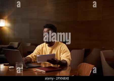 African American young businessman working on laptop with documents while sitting at table in restaurant Stock Photo
