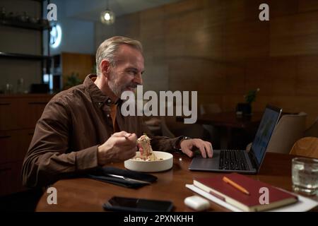Mature man using laptop while sitting at table and eating lunch in cafe Stock Photo