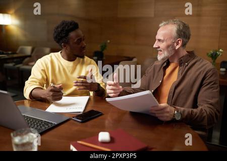 Two colleagues discussing contract together while sitting at table with laptop at meeting in cafe Stock Photo